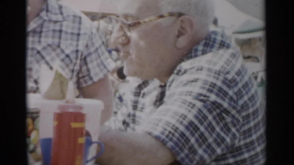 The older man sits at the picnic table in side view, surrounded by bottles, a tin, and standing guests in 1950s summer attire.