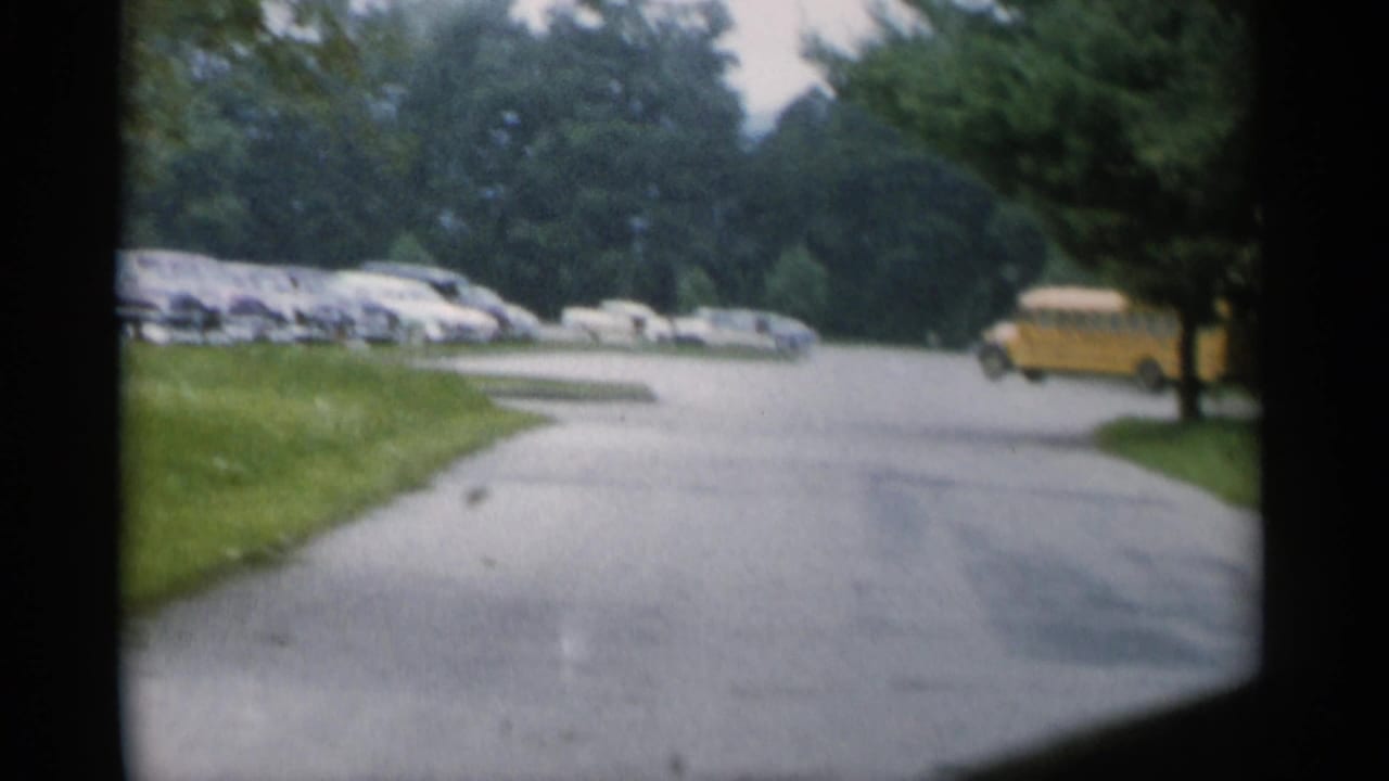 Paved road with parked classic cars, trees, and a distant yellow bus ahead.