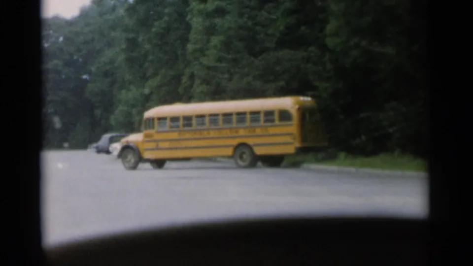 1956 Jones Beach frame with a yellow school bus moving across a paved road, seen through a vehicle windshield near wooded roadside parking.