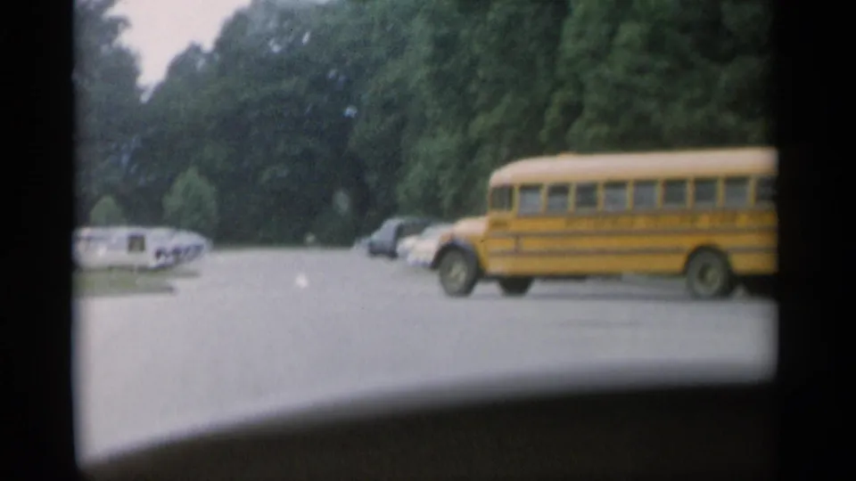 1956 Jones Beach home-movie frame showing a yellow school bus near the camera car, with classic parked cars and trees in the background.