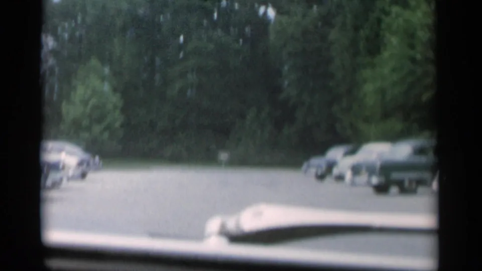 1956 Jones Beach frame looking over a car hood toward a paved road lined with parked classic cars and dense trees.