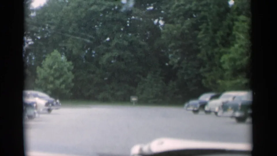 1956 Jones Beach view from inside a car, looking across a paved parking area with rows of midcentury automobiles and dense trees.