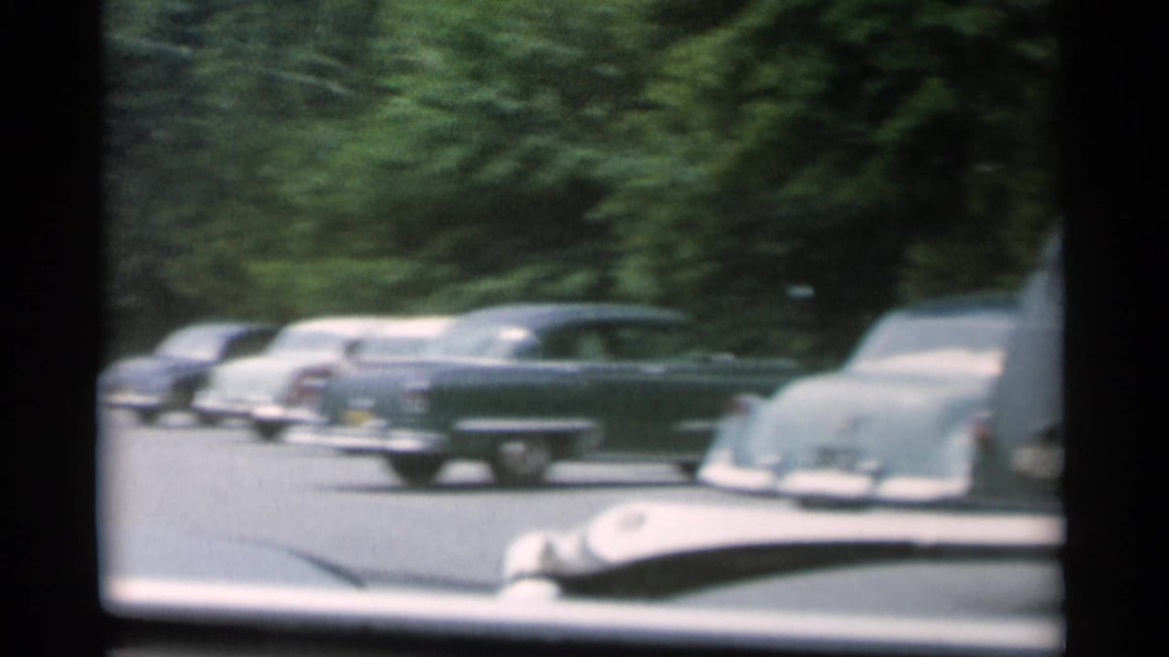 Parked cars and a car hood seen through a divided windshield or window frame.