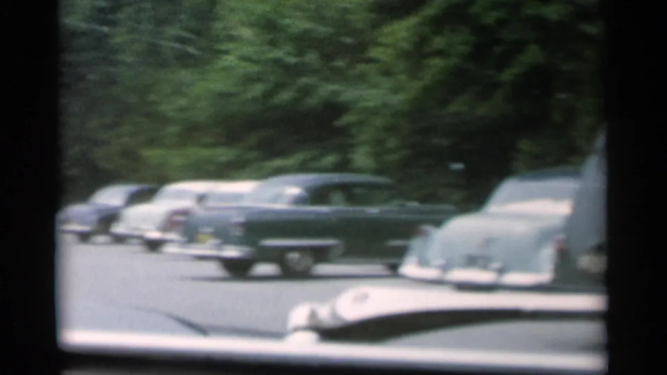 1956 Jones Beach parking-lot view with chrome-trimmed cars, a visible hood ornament area, and dark window framing from inside the vehicle.