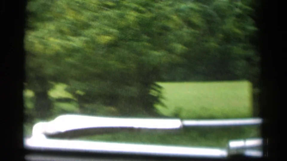 1956 Jones Beach home-movie frame from a moving car, with green shrubs, lawn, and the vehicle hood softened by motion blur.