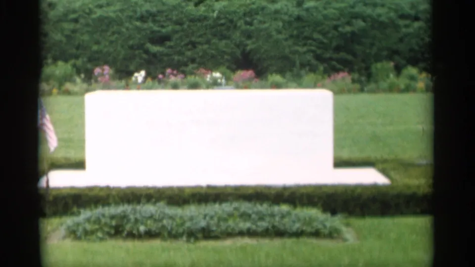 1956 Jones Beach frame showing a white grave monument on manicured grounds with hedges, flowers, and a small American flag nearby.