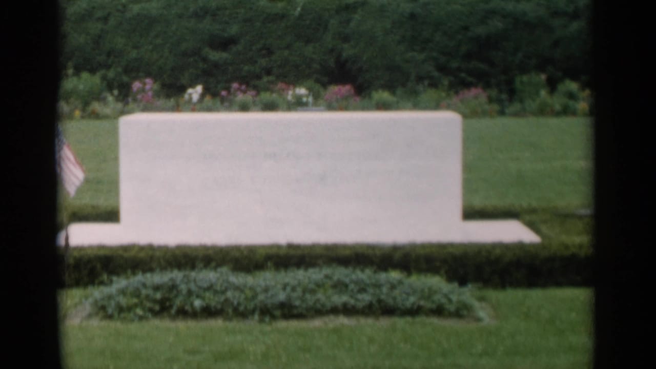 Pale rectangular stone feature beside lawn, hedges, flowers, and a small American flag.