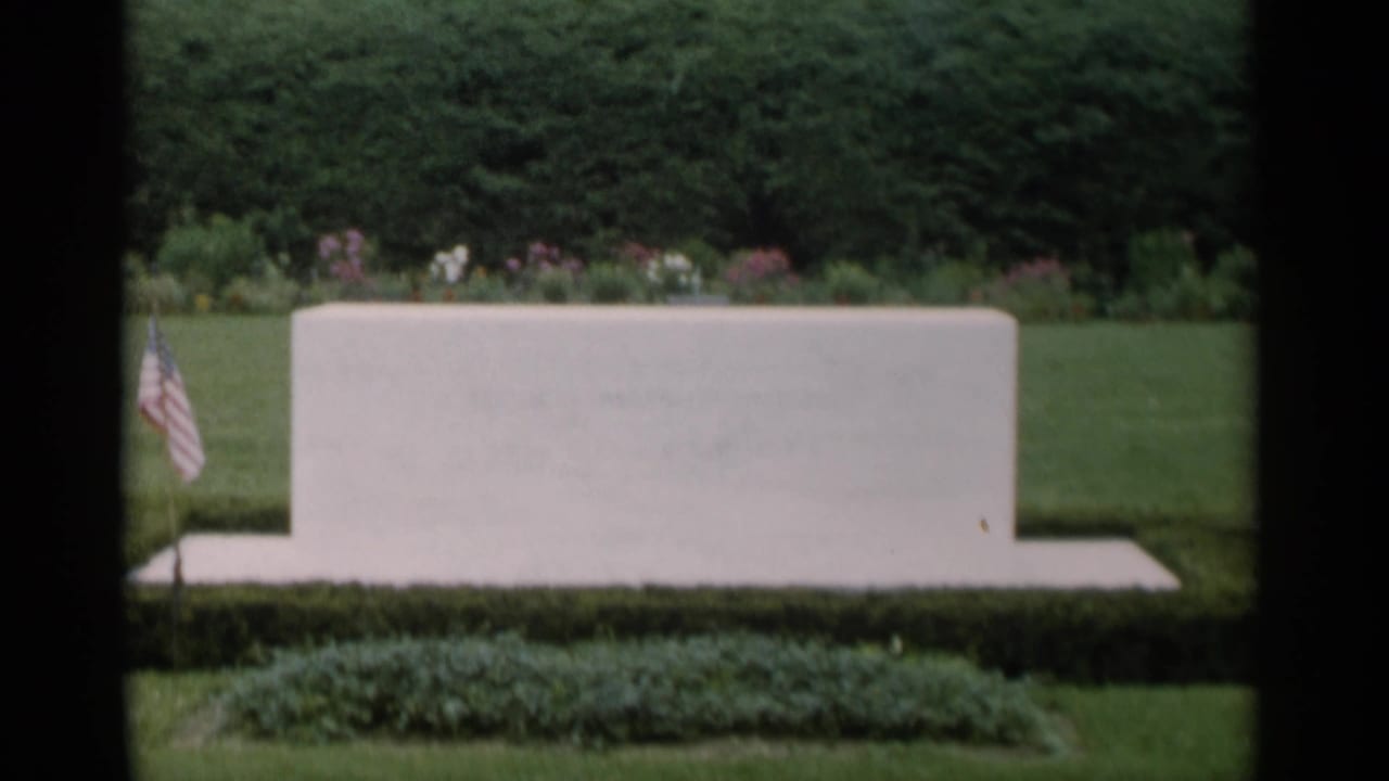 Stone feature centered in a manicured garden with lawn, hedges, flowers, and a small flag.