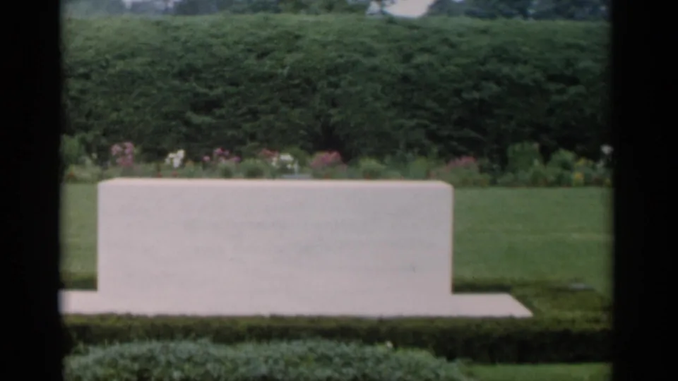 1956 home-movie frame at Jones Beach with a pale stone feature below a dense hedge and flowerbed, bordered by dark film edges.