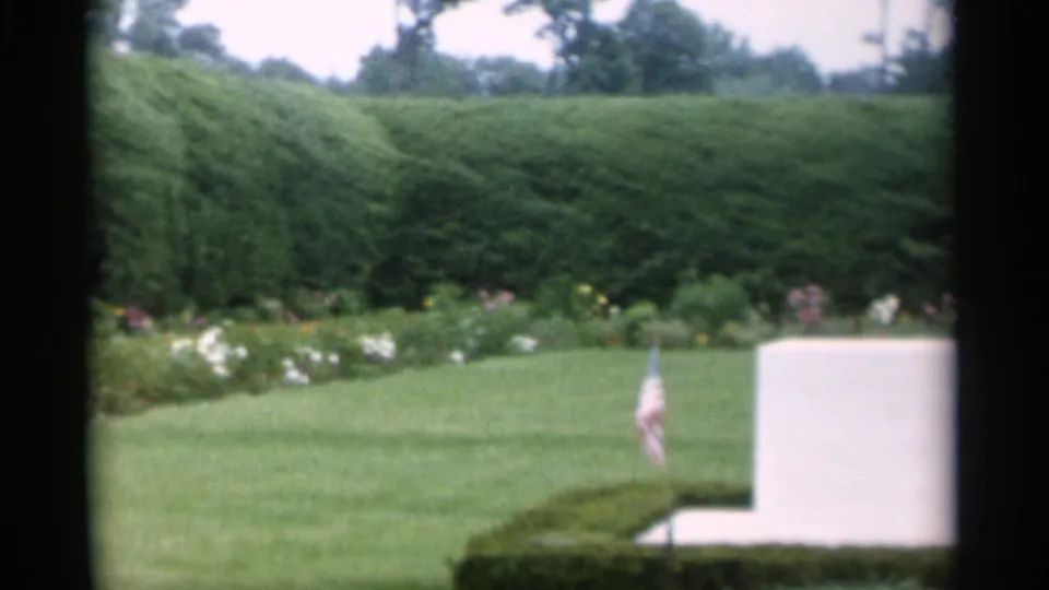 1956 Jones Beach home-movie frame showing a white memorial-like marker, small American flag, clipped hedges, and flower beds.