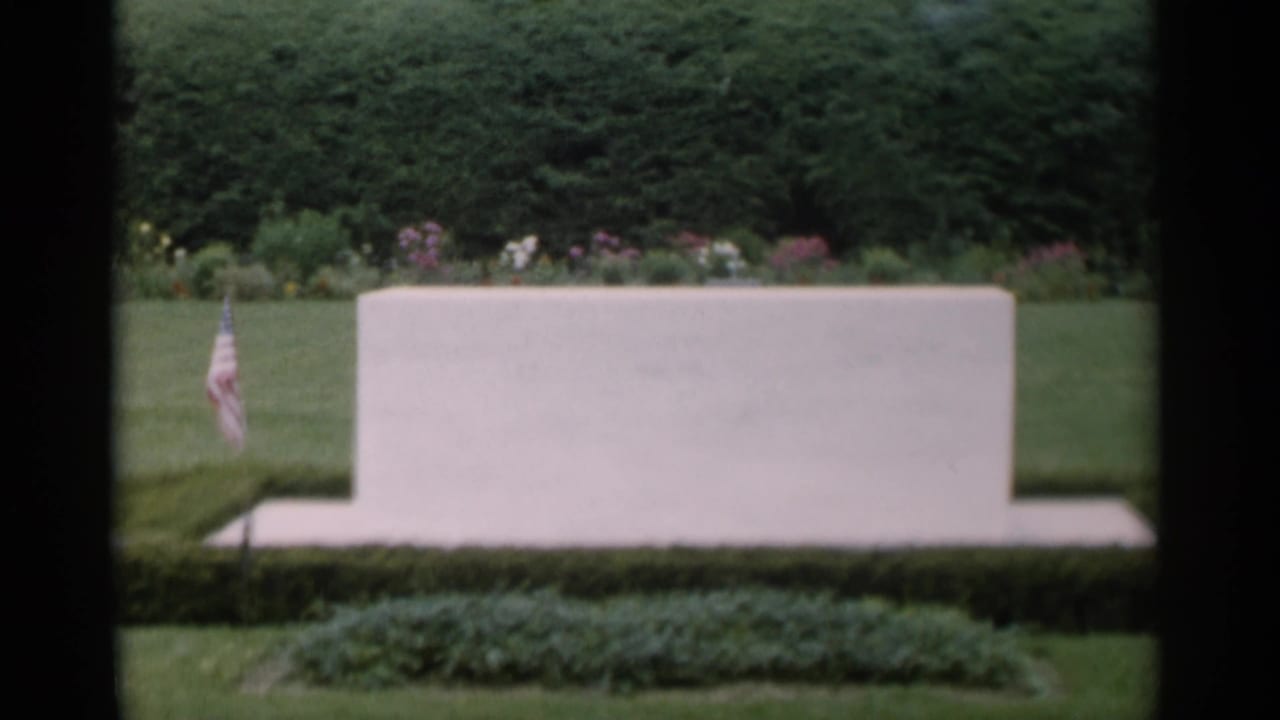 White stone marker beside hedges, flowers, lawn, and a small American flag.
