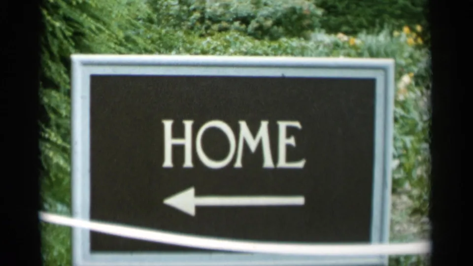 1956 Jones Beach home-movie frame showing a black HOME directional sign with a left arrow in front of leafy planting.