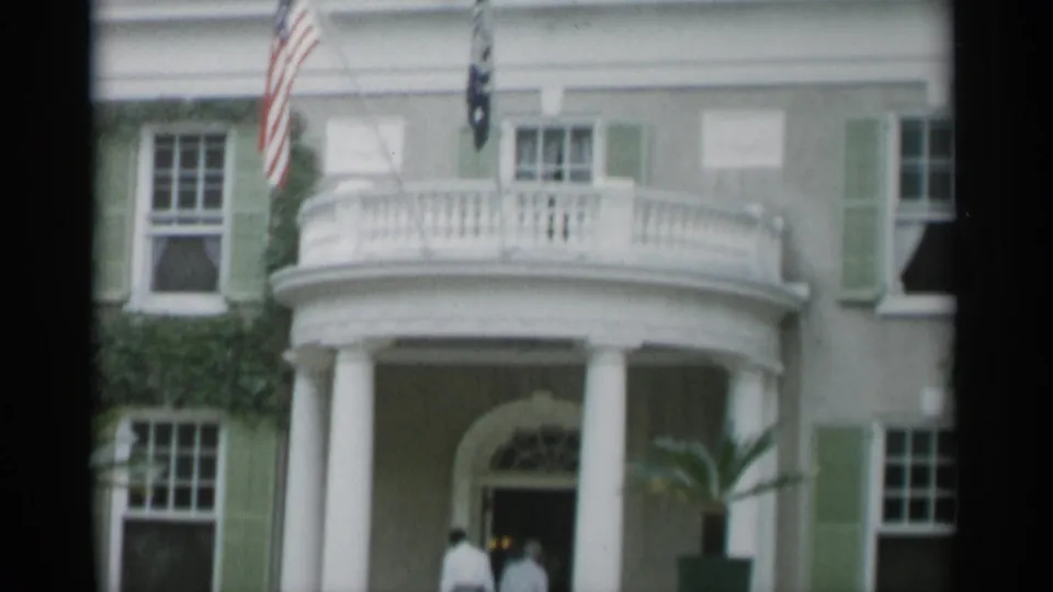 Home-movie frame from Jones Beach showing the formal facade, curved balcony, white columns, flags, and visitors near the entrance.