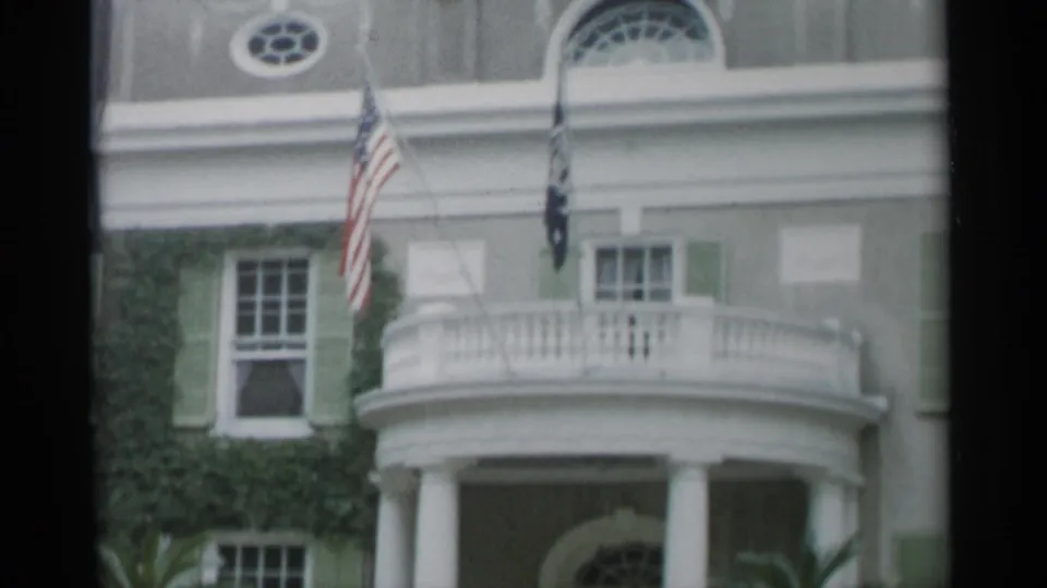 1956 Jones Beach frame showing the building's upper facade, flags, round window, green shutters, balcony, and portico below.