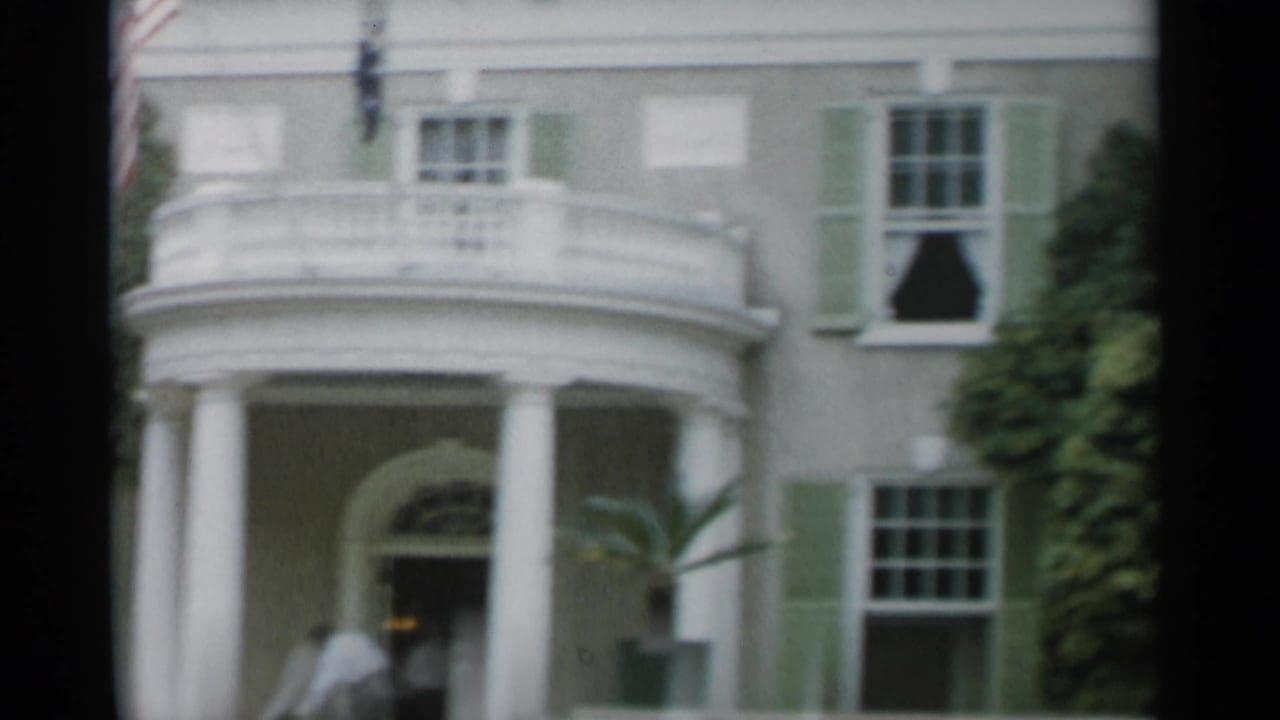 Columned portico with green shutters, balcony, doorway, and indistinct figures.