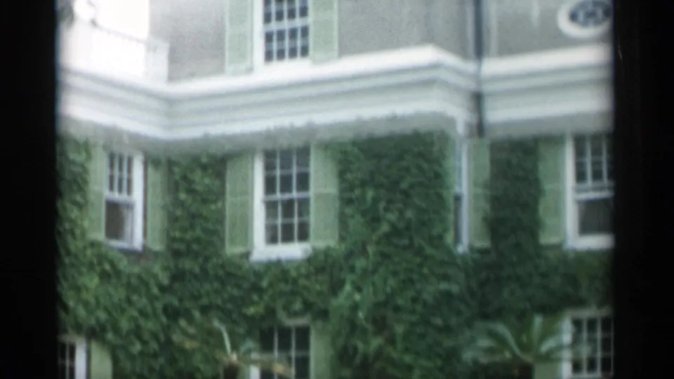 1956 Jones Beach frame showing the ivy-covered lower facade beneath a curved balcony, with sash windows, green shutters, and palm planting.