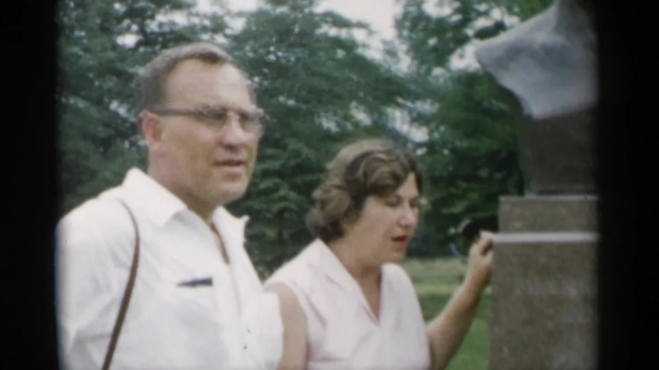 1956 Jones Beach home-movie frame showing an adult couple beside the Roosevelt monument, with trees and lawn in the background.
