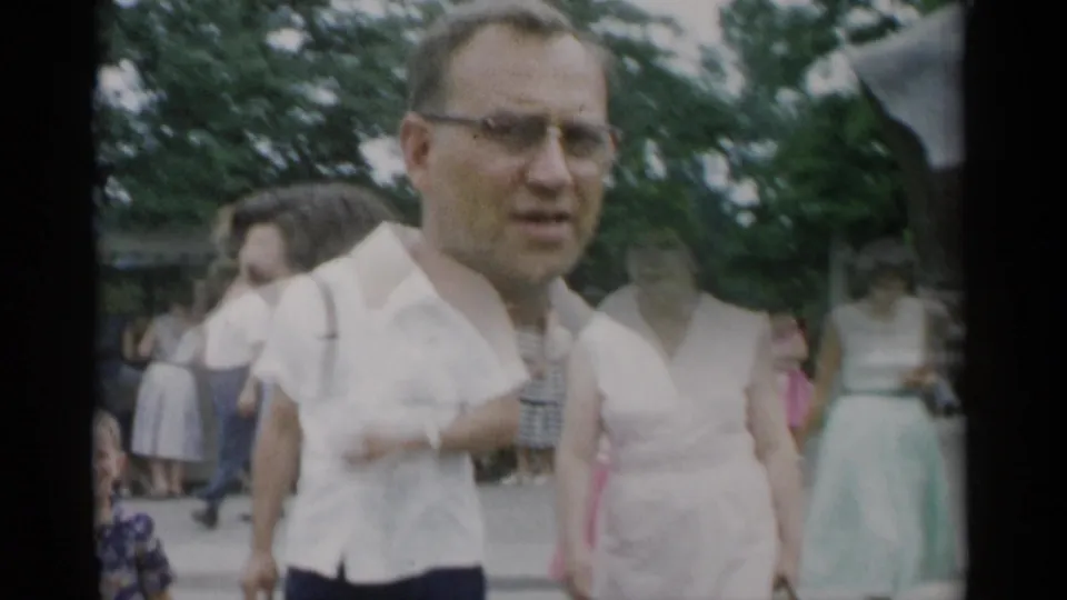 1956 Jones Beach frame with a man in glasses near the monument area, other visitors on a park walkway, and trees behind them.