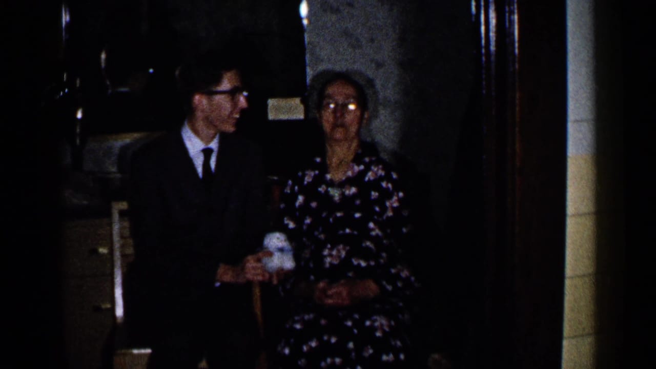 Young man in a dark suit sits beside an older woman in a floral dress in a dim interior.
