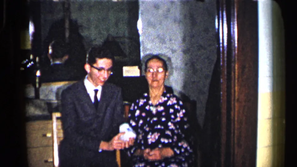 1960 Duluth frame of a suited young man holding a small white gift beside an older woman in a floral dress indoors.