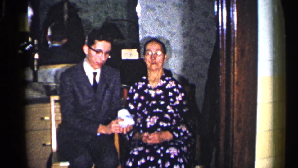 1960 Duluth home-movie still with two seated adults, the young man holding a small white gift in a dim private room.