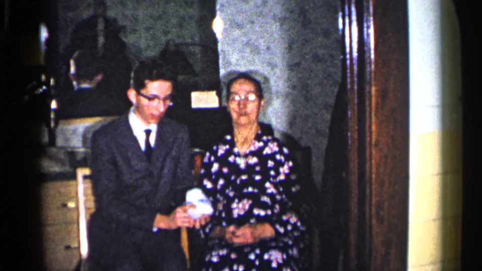1960 Duluth frame showing a young man in a suit and an older woman in a floral dress seated with a small gift indoors.