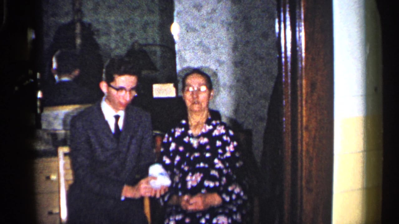 Young man in a dark suit stands beside an older woman seated in a living room.