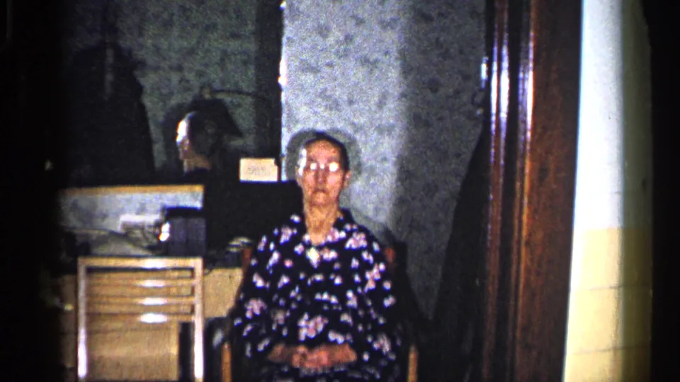 The camera frames an older woman seated in a chair in a Duluth living room, with a cabinet, dark object, wallpaper, and wood doorway behind her.