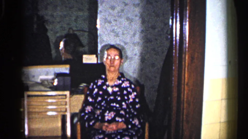 An older woman in glasses and a dark floral dress sits with hands clasped in a 1960 Duluth private interior, framed by wallpaper and wood trim.