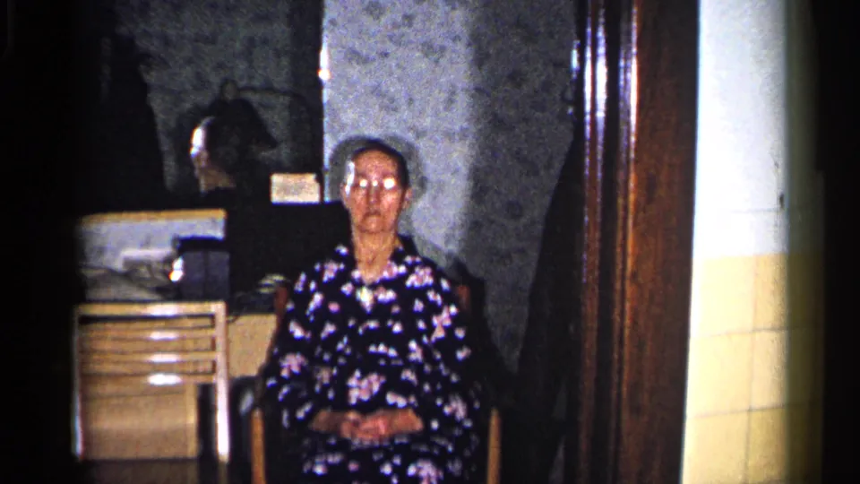 The final frame holds on the older woman seated in a dim Duluth living room, with reflective glasses, floral dress, cabinet drawers, and wood doorway visible.