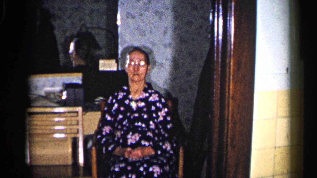 In a 1960 Duluth home-movie frame, an elderly woman in a dark floral dress sits indoors beside a dresser or cabinet.