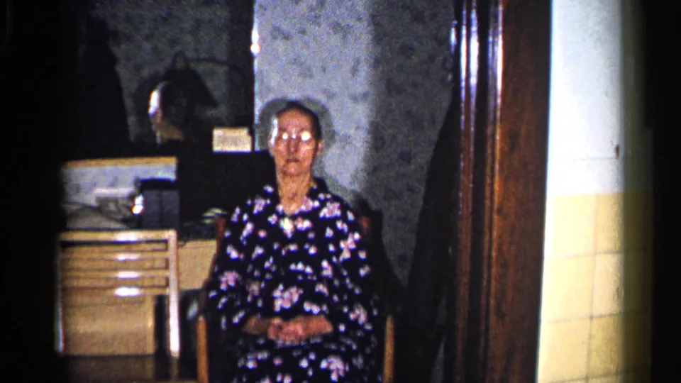 In a 1960 Duluth home-movie frame, an elderly woman in a dark floral dress sits indoors beside a dresser or cabinet.