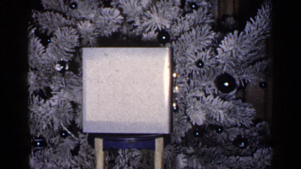 A final close frame shows the white gift box in front of the flocked Christmas tree, with dark ornaments visible around it.