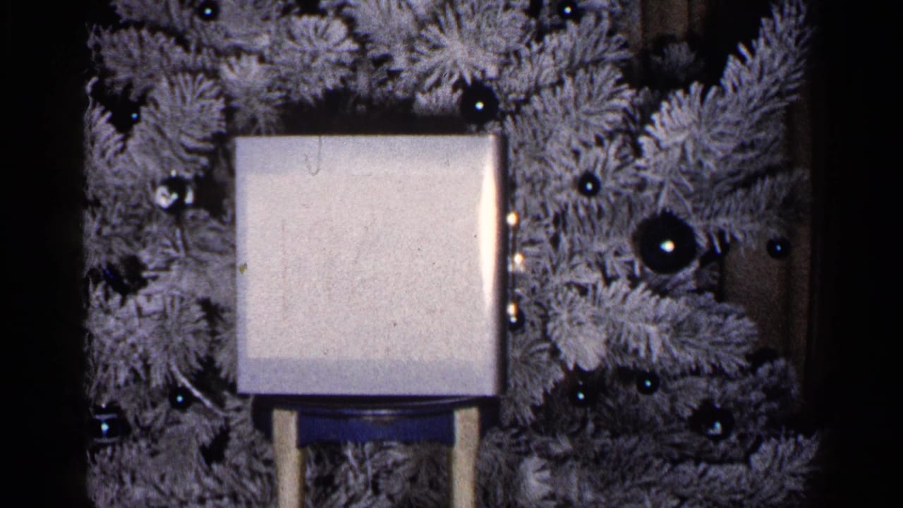 1960 Duluth home-movie frame showing a pale square object on a stand before a flocked Christmas tree with dark reflective ornaments.