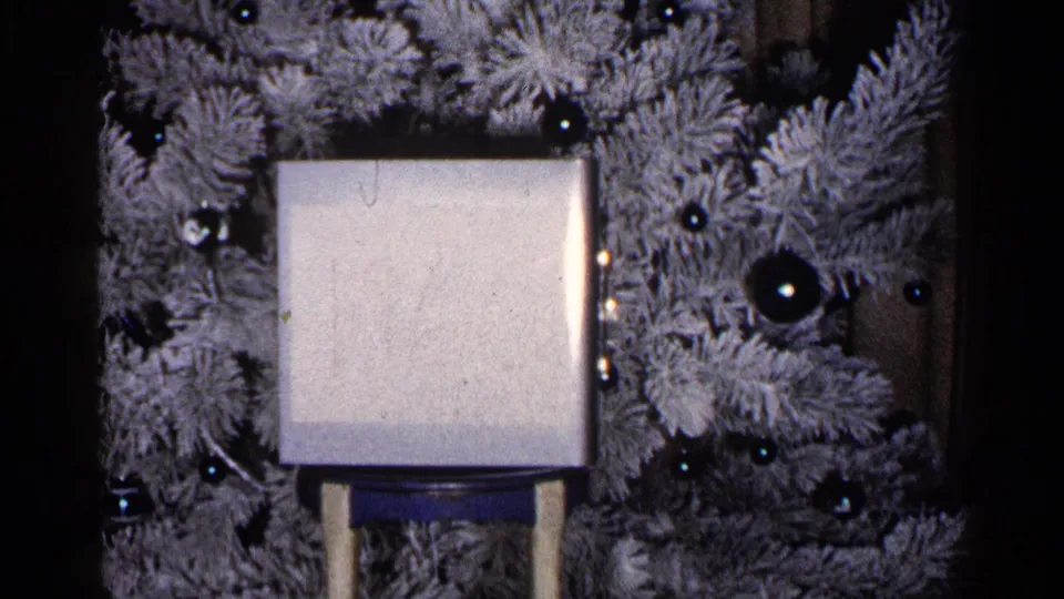 1960 Duluth home-movie frame showing a pale square object on a stand before a flocked Christmas tree with dark reflective ornaments.