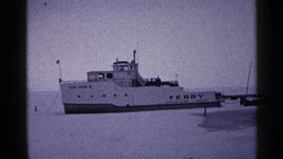 1960 Duluth frame of a white ferry marked FERRY, motionless on frozen lake ice with dock structures nearby.