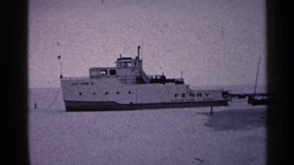 1960 Duluth home-movie view of the ferry broadside on frozen water, with side lettering and nearby mooring elements visible.