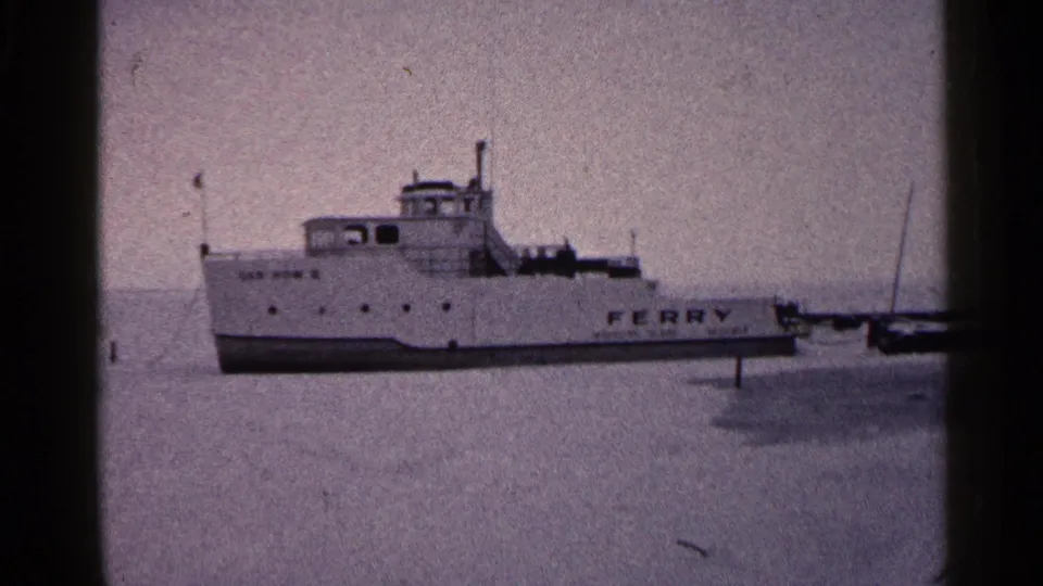 1960 Duluth home-movie frame showing a white ferry anchored on a frozen lake with muted 8mm color and dark frame edges.