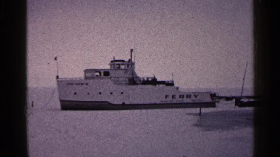 1960 Duluth 8mm frame of a white ferry seen broadside on frozen water, with the word FERRY readable on the hull.