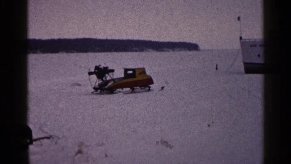 1960 Duluth 8mm frame showing a yellow-red snowmobile resting on lake ice, with shoreline and part of the ferry in the background.