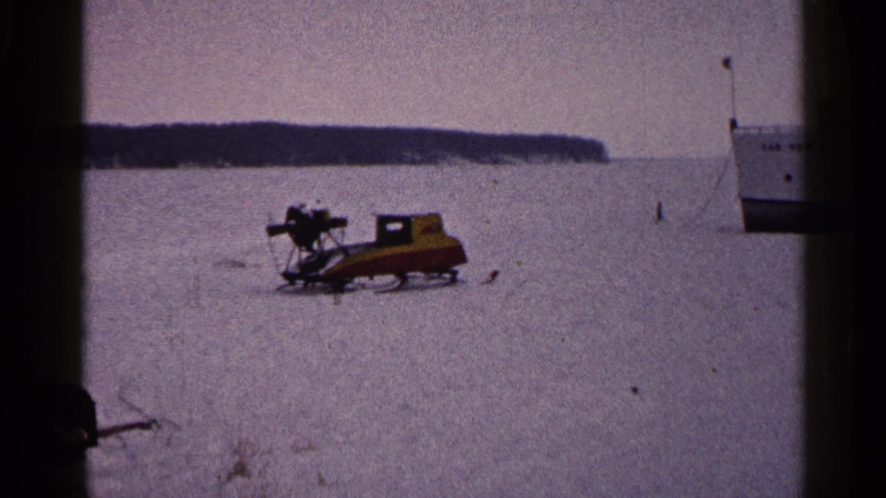 Circa-1960 Duluth winter frame showing a yellow and red snowmobile on frozen lake ice near a white boat.