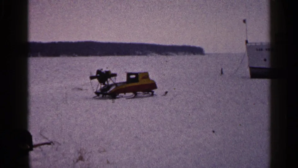 Circa-1960 Duluth winter frame showing a yellow and red snowmobile on frozen lake ice near a white boat.