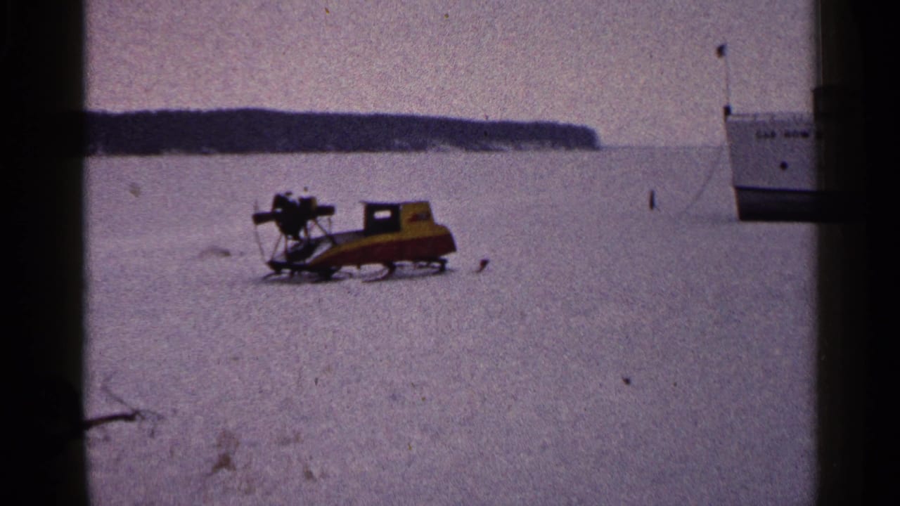 A snowmobile sits or moves slowly across frozen lake ice in Duluth, Minnesota, with a white boat nearby.