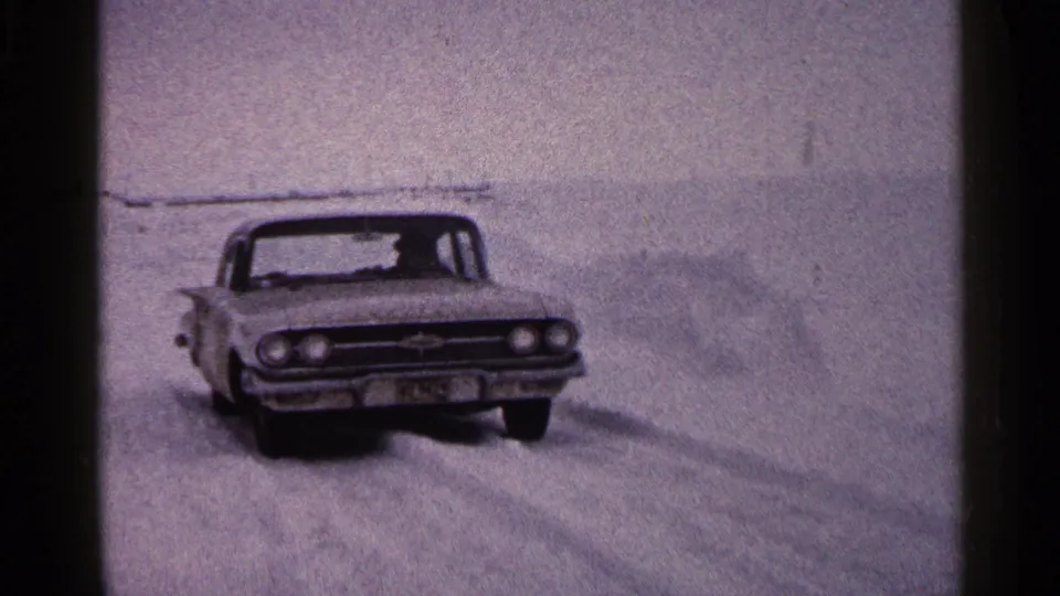 The sedan continues across the frozen lake in Duluth, with the driver faintly visible through the windshield.