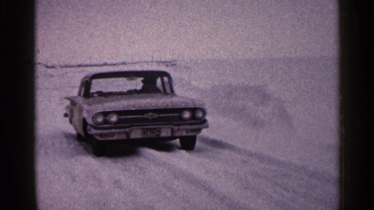 1960 Duluth home-movie frame showing a classic car pushing through deep snow on open winter ground.
