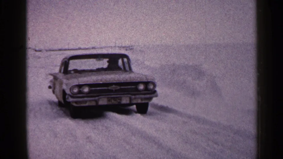 1960 Duluth home-movie frame showing a classic car pushing through deep snow on open winter ground.