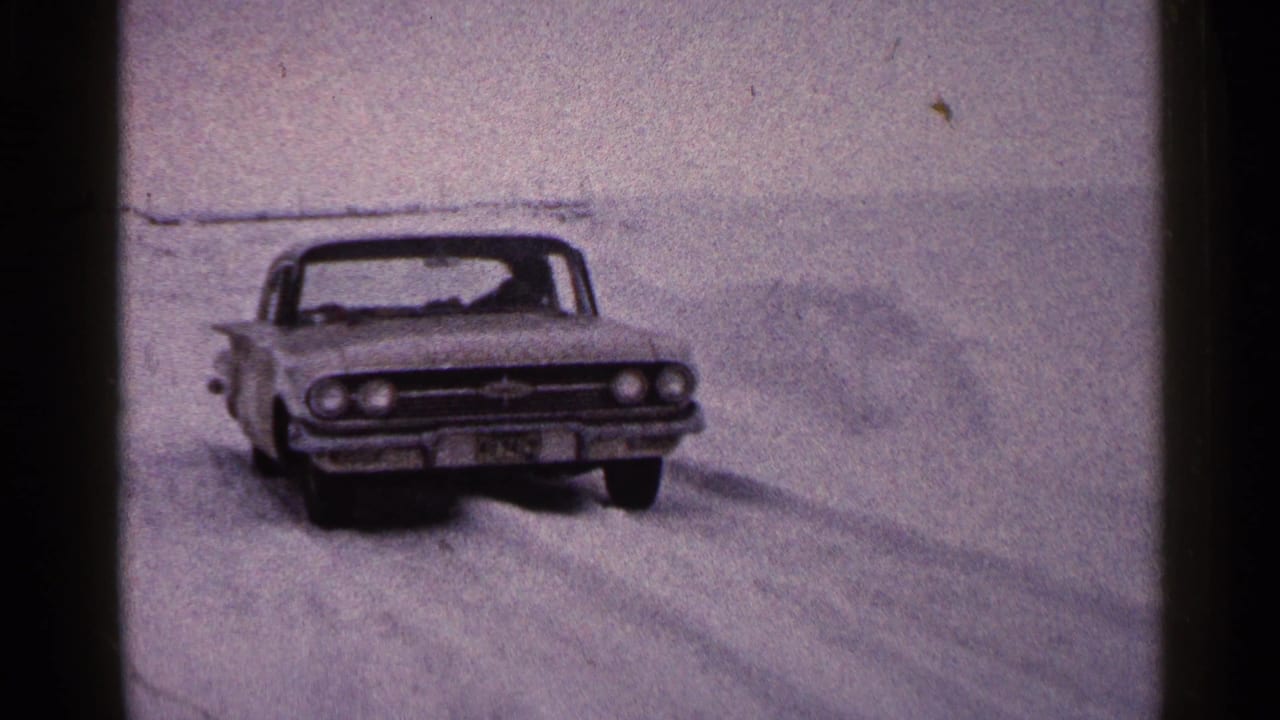 1960 Duluth winter frame with a classic car angled toward camera amid deep snow and soft 8mm grain.