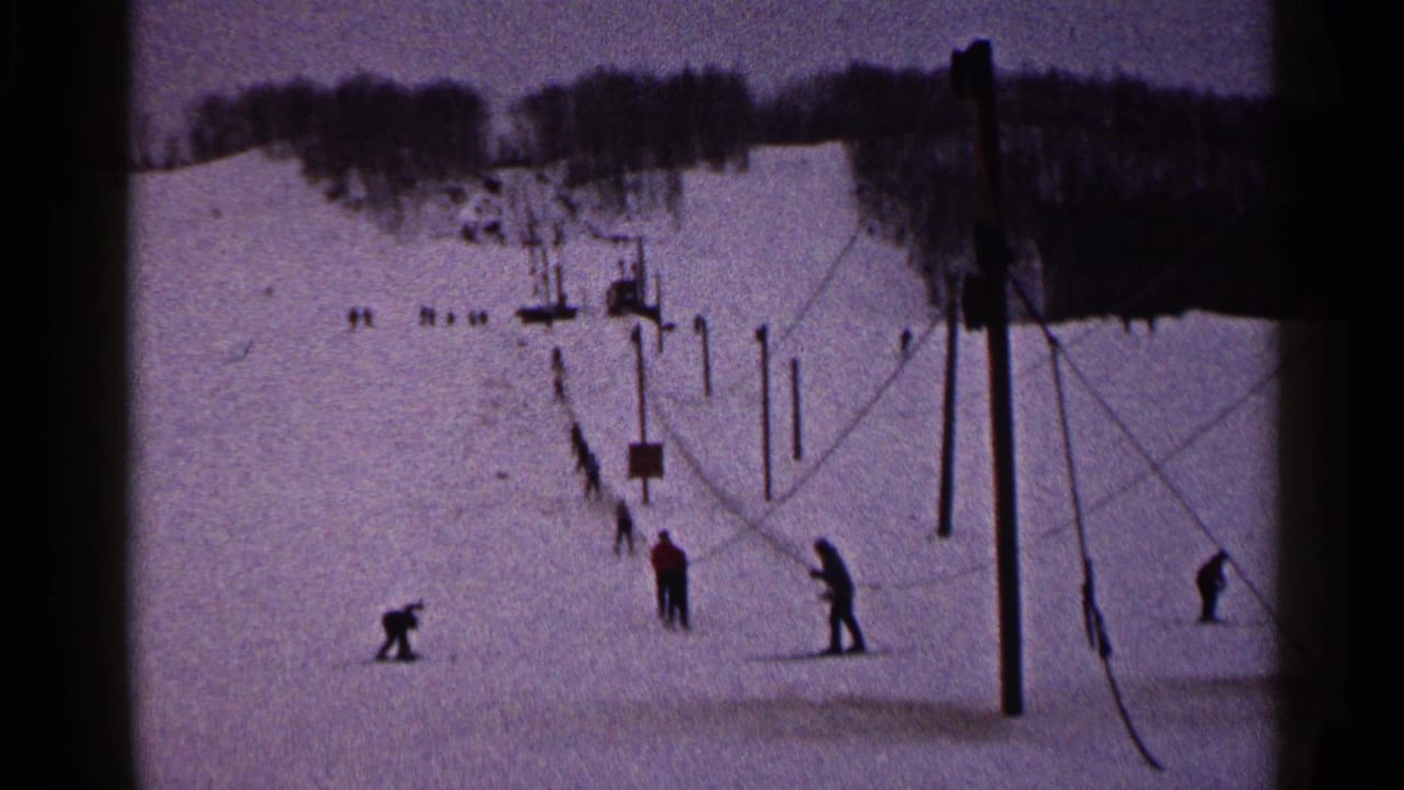 1960 Duluth home-movie frame showing distant skiers on a snowy slope with lift poles and a wooded ridge.