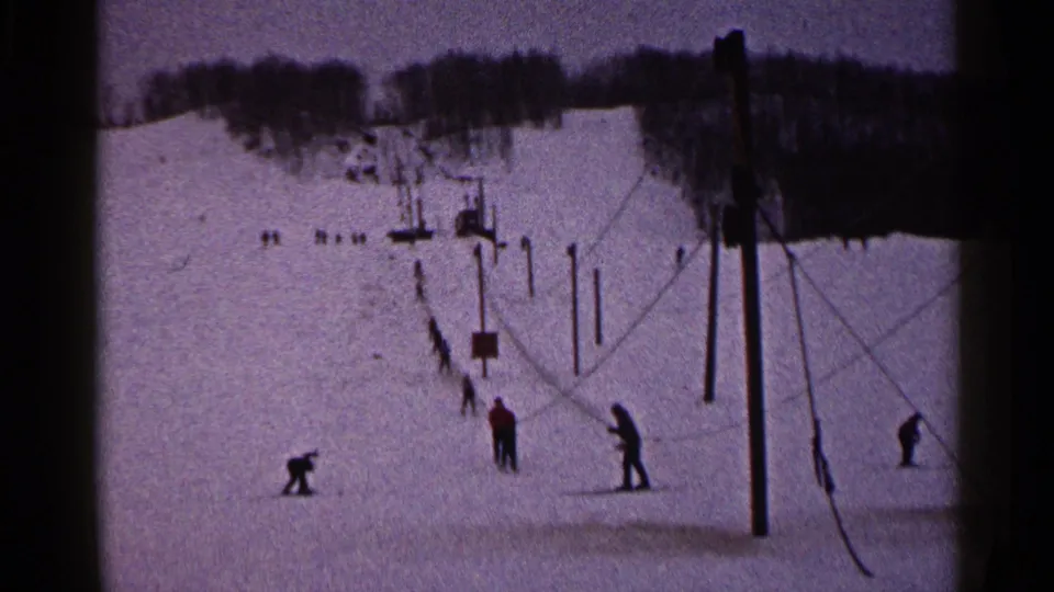 1960 Duluth home-movie frame showing distant skiers on a snowy slope with lift poles and a wooded ridge.