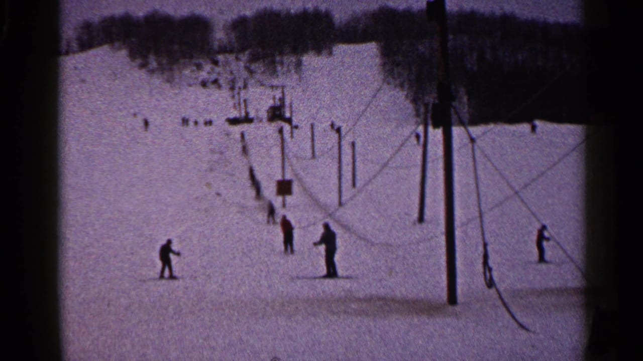 1960 Duluth winter frame with several distant skiers, lift poles, cables, and a wooded ridge above the slope.